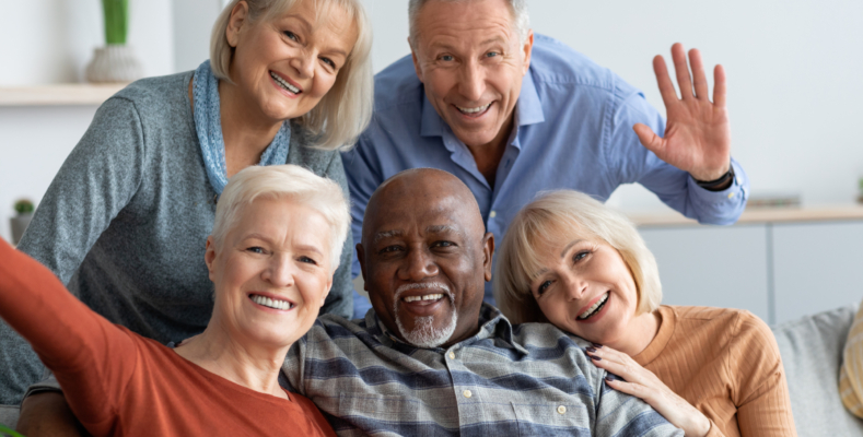 Multiracial group of happy positive elderly people men and women in casual outfits taking selfie together while chilling together, hugging and smiling at camera, nursing home interior